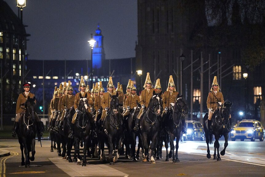 People in royal military uniforms ride on horseback through a dark street