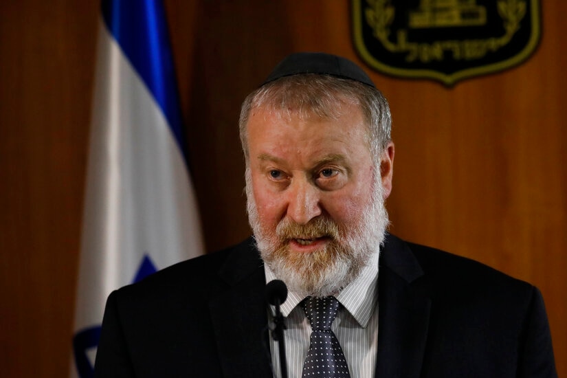 A man with a white beard wears a kippah and stands in front of two Israeli flags while speaking on a lectern.