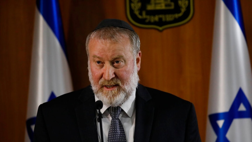 A man with a white beard wears a kippah and stands in front of two Israeli flags while speaking on a lectern.