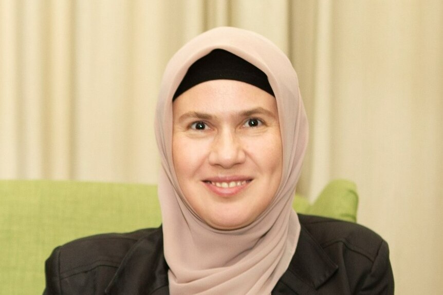 A photo of a woman wearing a beige hijab and black under cap. She is smiling. 