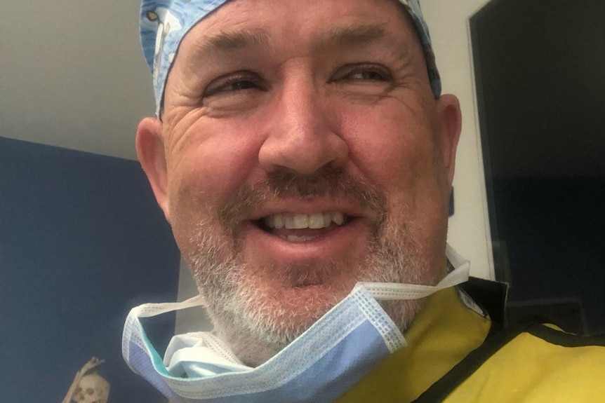 A doctor smiling and wearing yellow scrubs and a mask.