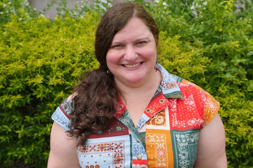 A woman with long brown hair and a colourful shirt smiles at the camera