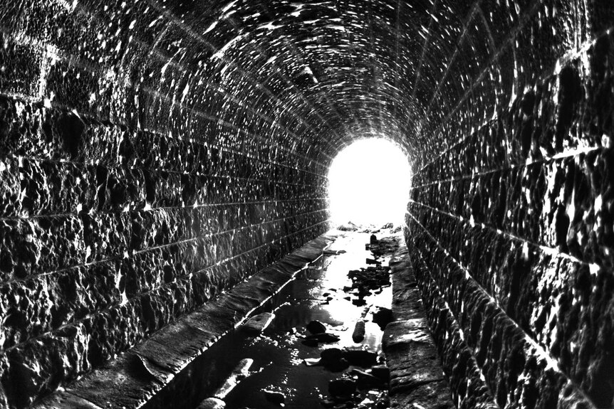 A black and white shot of an old tunnel with old sandstone-like bricks leading to light at the end of the tunnel