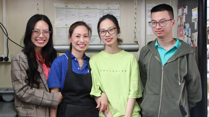 A Vietnamese family of four stand in a commercial kitchen with big smiles on their faces.