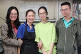 A Vietnamese family of four stand in a commercial kitchen with big smiles on their faces.