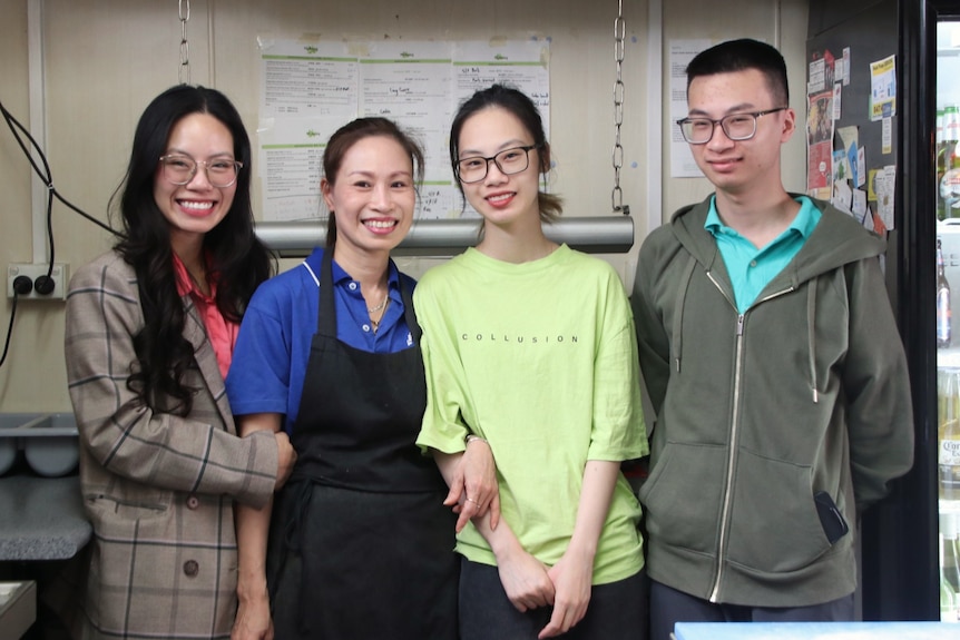 A Vietnamese family of four stand in a commercial kitchen with big smiles on their faces.