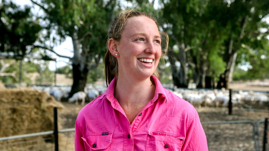Woman with red hair wearing pink shirt smiles with sheep, trees and hay bales visible in background