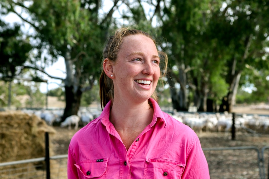 Woman with red hair wearing pink shirt smiles with sheep, trees and hay bales visible in background