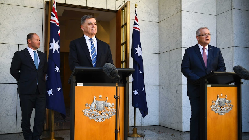 Brendan Murphy and Scott Morrison stand at podiums. Josh Frydenberg stands behind them besides Australian flags