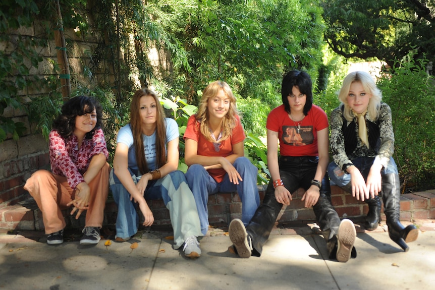 Five young women with 1970s style outfits and haircuts sit on low to ground brick wall near green forest area on sunny day.