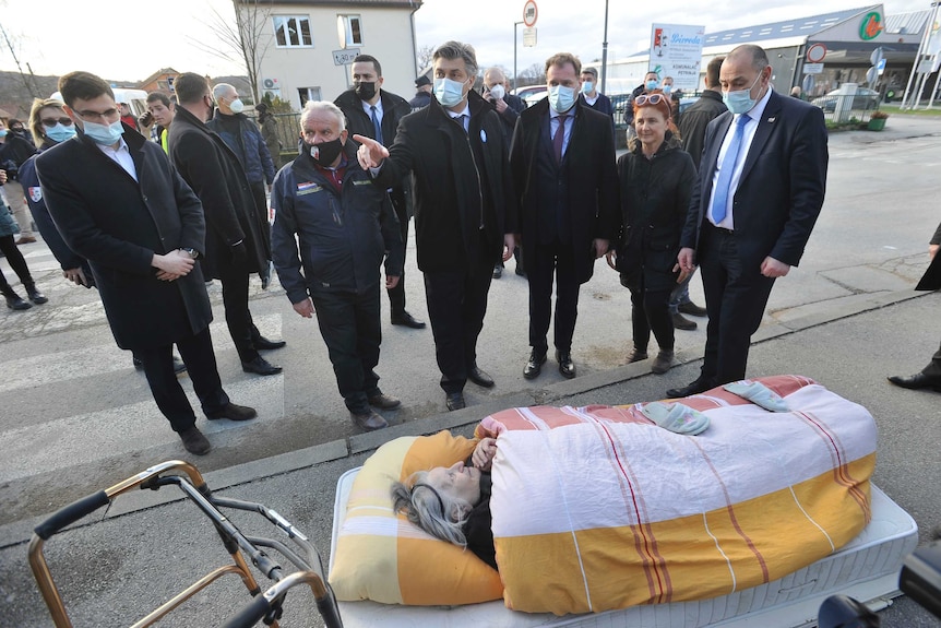 A group of men and women in face masks star at an old lady in a stretcher on the street.