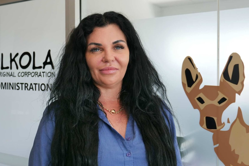 Woman with long, dark hair standing in front of an office with "Olkola Aboriginal Corporation" sign and picture of dingo.