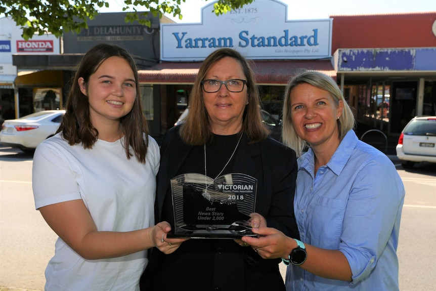 Three women stand in a line with an award