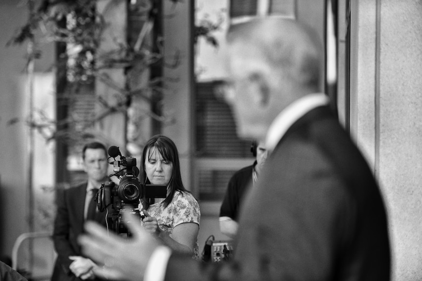Black and white photo of woman operating a camera pointed at blurred profile of Scott Morrison.
