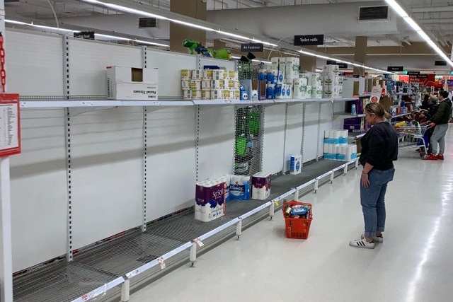 A woman stands looking at mostly empty shelves, where a handful of packs of toilet paper sit.