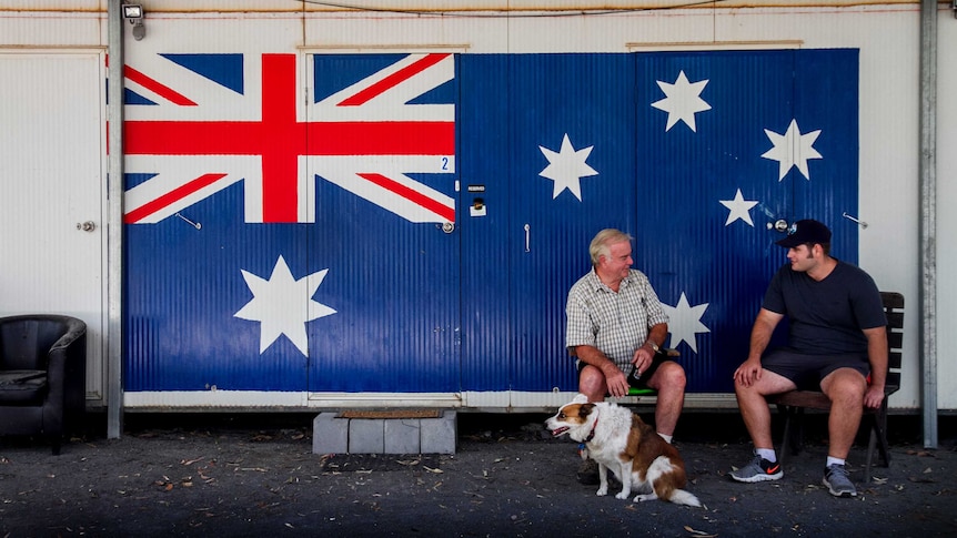 Two men and a dog sit in front of a donga with a large Australian Flag painted on it.