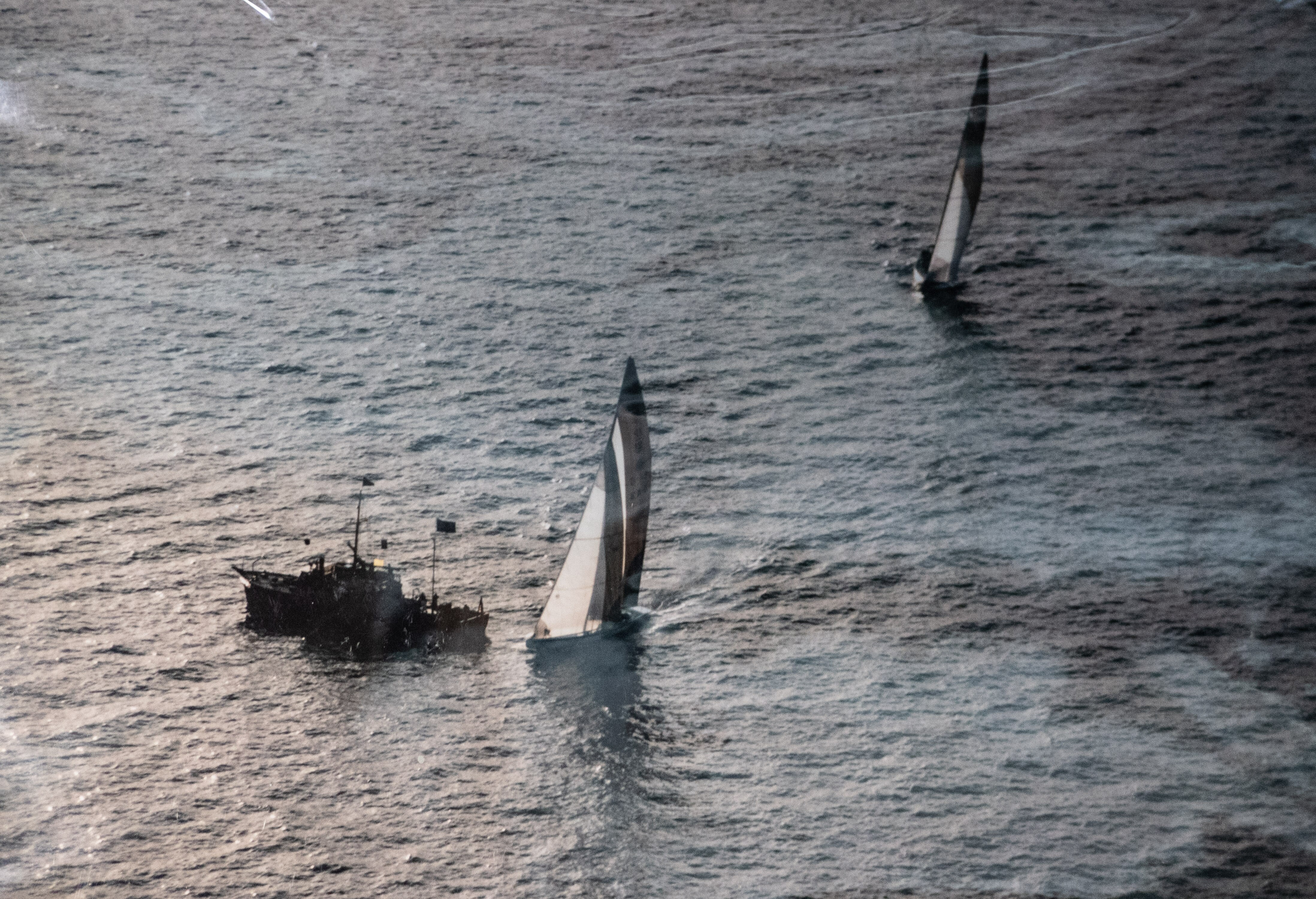 A yacht passes smaller observation boats. It is trailed by another racing vessel.