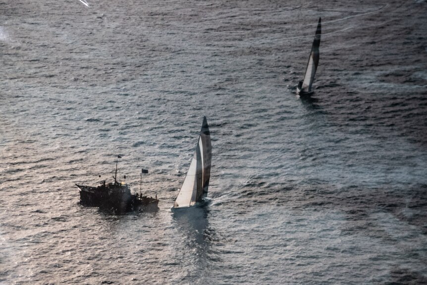 A yacht passes smaller observation boats. It is trailed by another racing vessel.