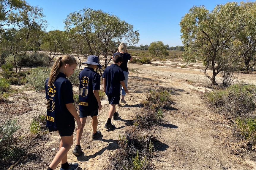 A woman and three children walk in a line along a dry, cracked environment. There are some trees and shrubs either side.