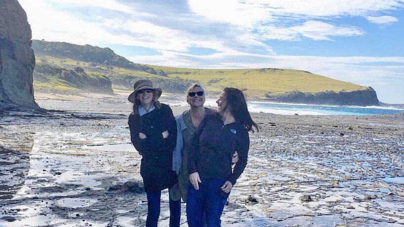Three women stand near a rugged coastline smiling at the camera.