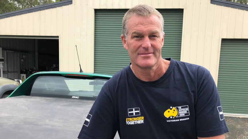 A smiling middle-aged man in a blue t-shirt leans on the bonnet of a car