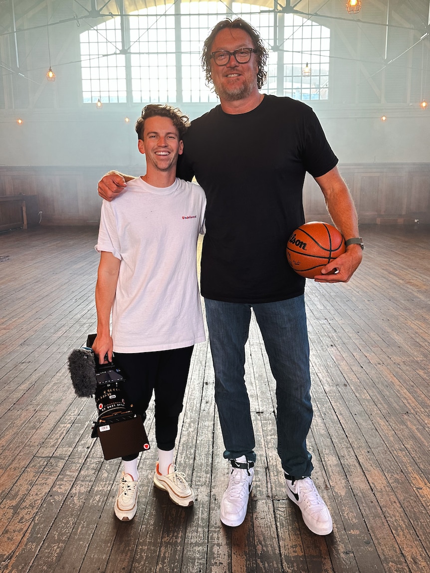 Sam Tolhurst and Luc Longley stand next to each other. Sam is holding a video camera and Luc holds a basketball.