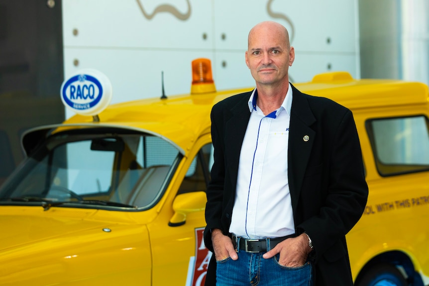 An image of Andrew Kirk standing in front of a yellow RACQ vehicle, with hands in his pockets, wearing a blue shirt and jacket.