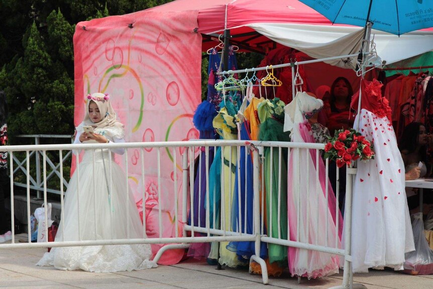 A woman in a hijab and wedding dress plays with her phone next to a market stall.