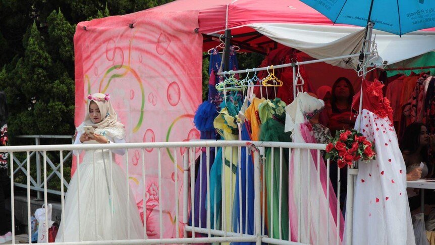 A woman in a hijab and wedding dress plays with her phone next to a market stall.