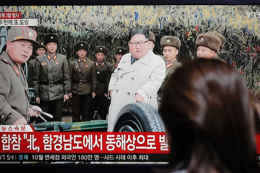 Kim Jong Un appears on a TV screen wearing a white jacket surrounded by soldiers.