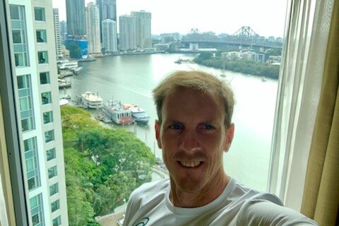 A man with blonde hair takes a selfie in front of a hotel window with a river, buildings and Brisbane's Storey Bridge in view
