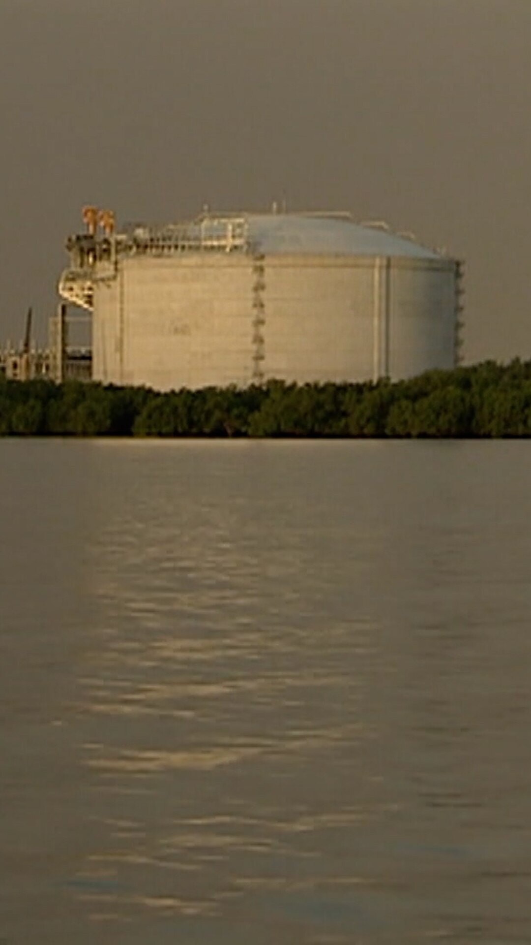 A large tank sits between a grey sky in the background and a muted body of water in the foregound
