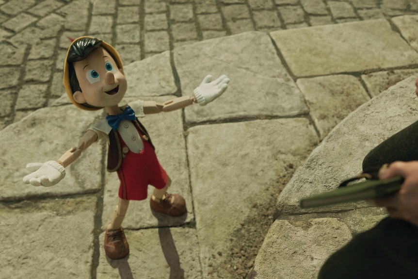 Overhead shot of humanoid toy, a boy wearing red and brown overalls, a cream hat and white gloves with his hands arms spread.