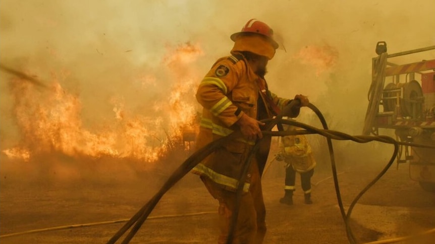 Two firefighters wrestle with a hose as a large grass fire burns behind them, sending smoke into the air.