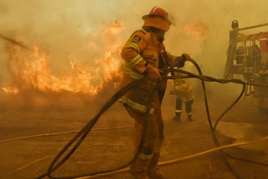 Two firefighters wrestle with a hose as a large grass fire burns behind them, sending smoke into the air.