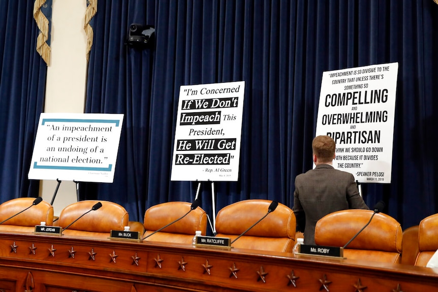 A large wooden desk with orange leather seats sit in front of three large placards detailing quotes critical of impeachment.