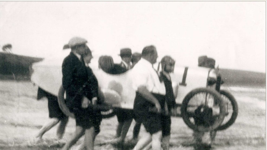 Nine men carry a vintage car across the beach
