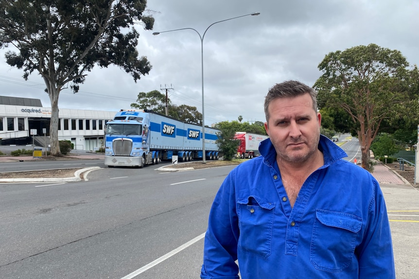 A man wearing a blue button up shirt with a concerned expression, behind him is a large blue truck in the middle of road