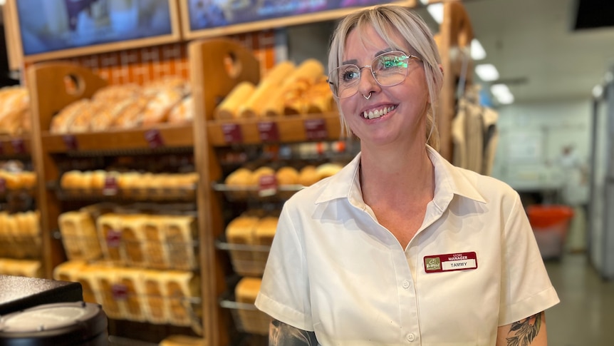 A young woman with blonde hair and glasses stands in front of rows of bread and smiles