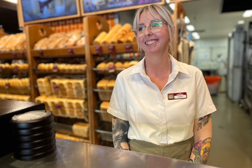 A young woman with blonde hair and glasses stands in front of rows of bread and smiles