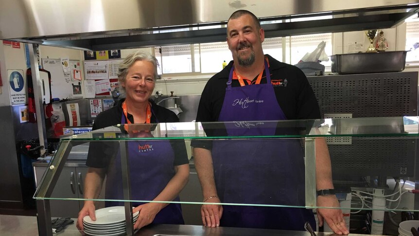 Two staff members wearing purple aprons stand behind a bain-marie at a homeless centre
