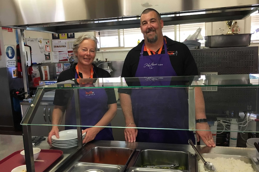 Two staff members wearing purple aprons stand behind a bain-marie at a homeless centre