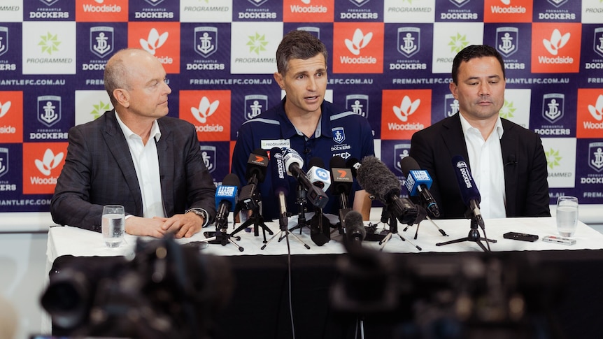Justin Longmuir speaks into microphones flanked by Dale Alcock and Peter Bell at a Dockers media conference indoors.