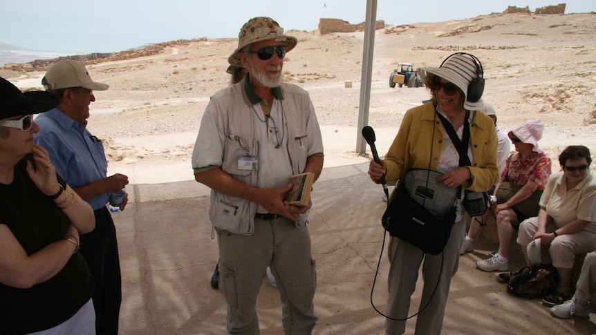 Kohn holding microphone and wearing headphones interviewing man in desert with people watching on.