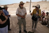 Kohn holding microphone and wearing headphones interviewing man in desert with people watching on.