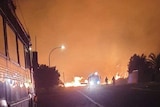 A fire truck on a road at night with a fire behind it, unidentified people on the street and a lot of smoke in the air.
