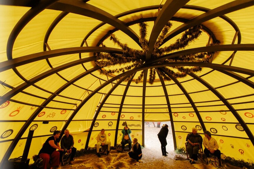 The inside of a dome with yellow canvas and wooden beams, possum skins on the floor and people sitting in chairs around