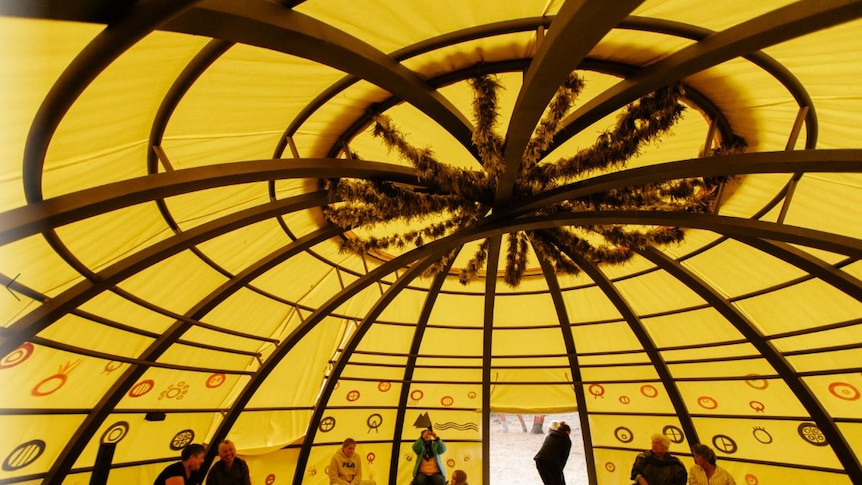 The inside of a dome with yellow canvas and wooden beams, possum skins on the floor and people sitting in chairs around
