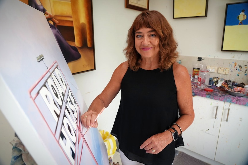 A smiling woman holds a paintbrush against a canvas in a studio