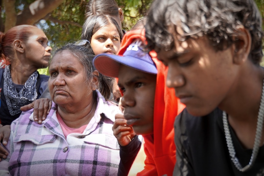 A group of Indigenous family members gathered in a park looking sombre and embracing each other